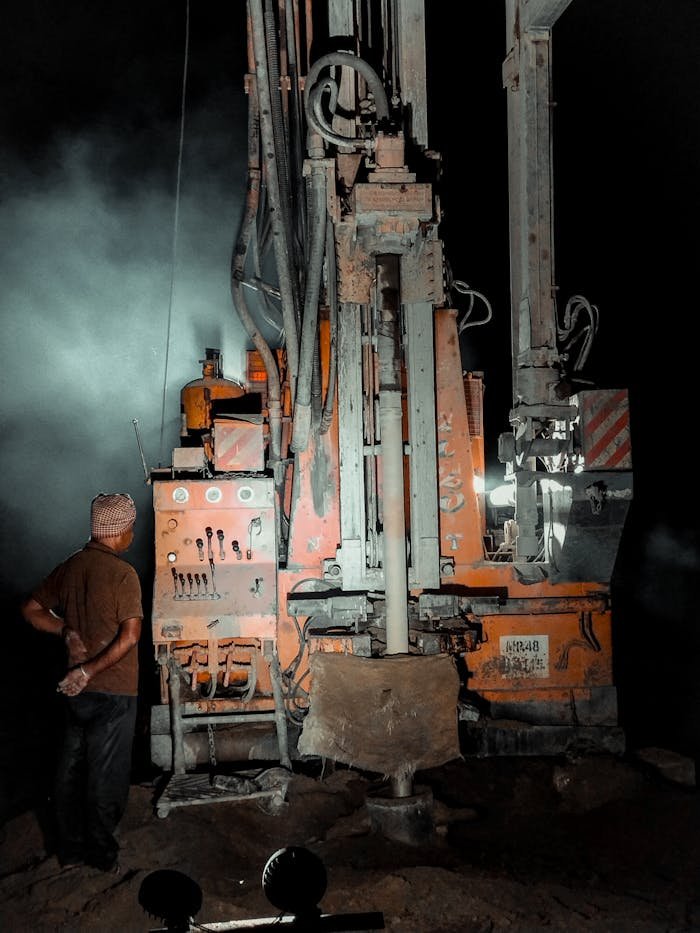 About A worker operates a water well drilling rig under illumination during nighttime.